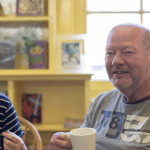 An older man smiles while holding a mug and an older woman smiles at him.