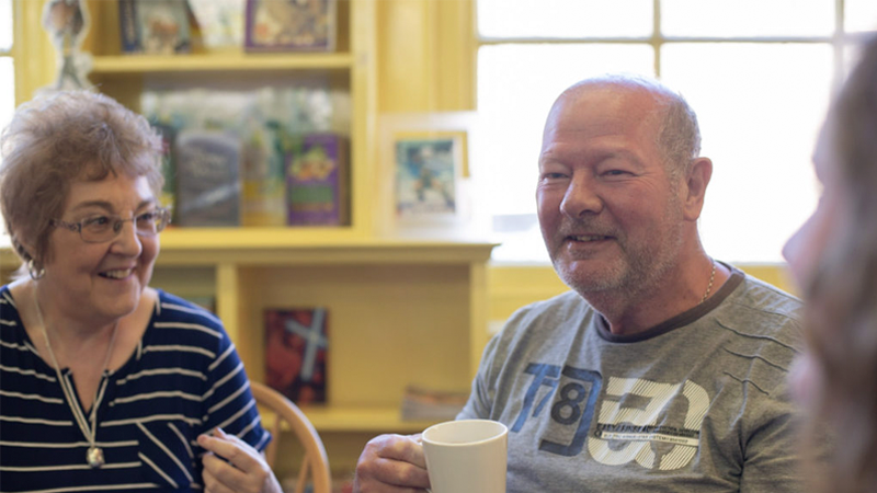 An older man smiles while holding a mug and an older woman smiles at him.