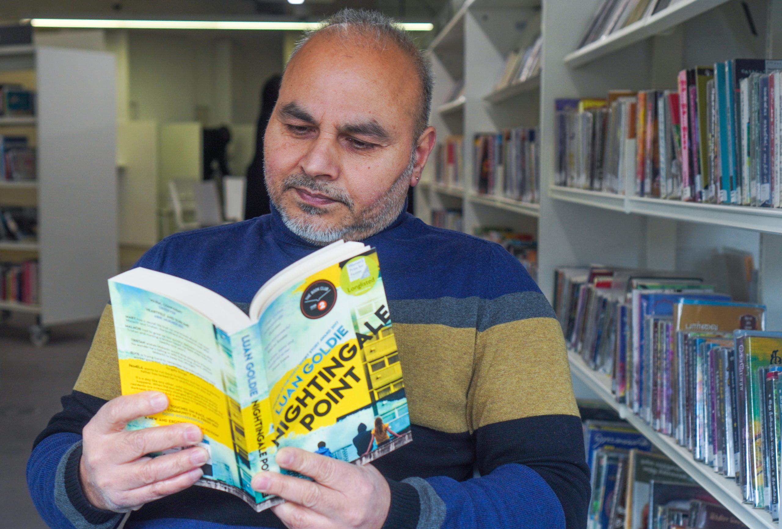 A middle aged man in a library smiles reading a copy of Nightingale Point by Luann Goldie 