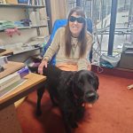 Photo of a woman wearing dark glasses smiling at the camera. She has her hand on her service dog.