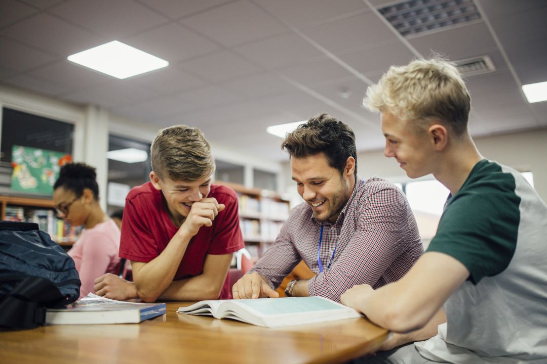 Photo of two teenage boys sat with an adult man looking at a book. There are bookshelves in the background.