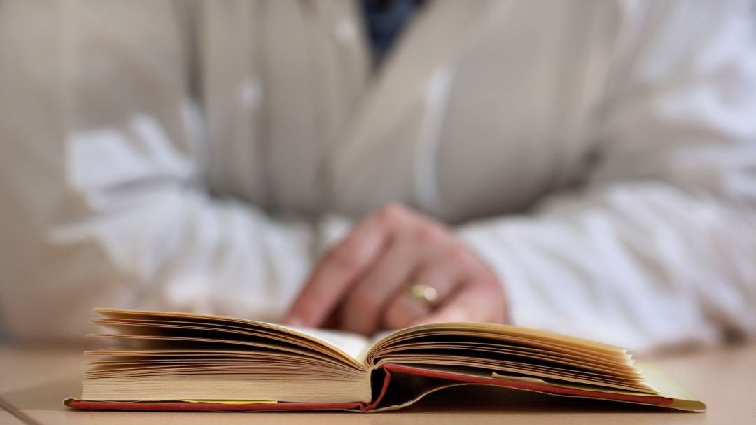 Photo of a person wearing a white lab coat and reading a book on a table.