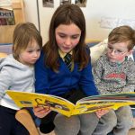 Photo of three children reading a book. The middle child is wearing school uniform.