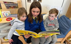 Photo of three children reading a book. The middle child is wearing school uniform.