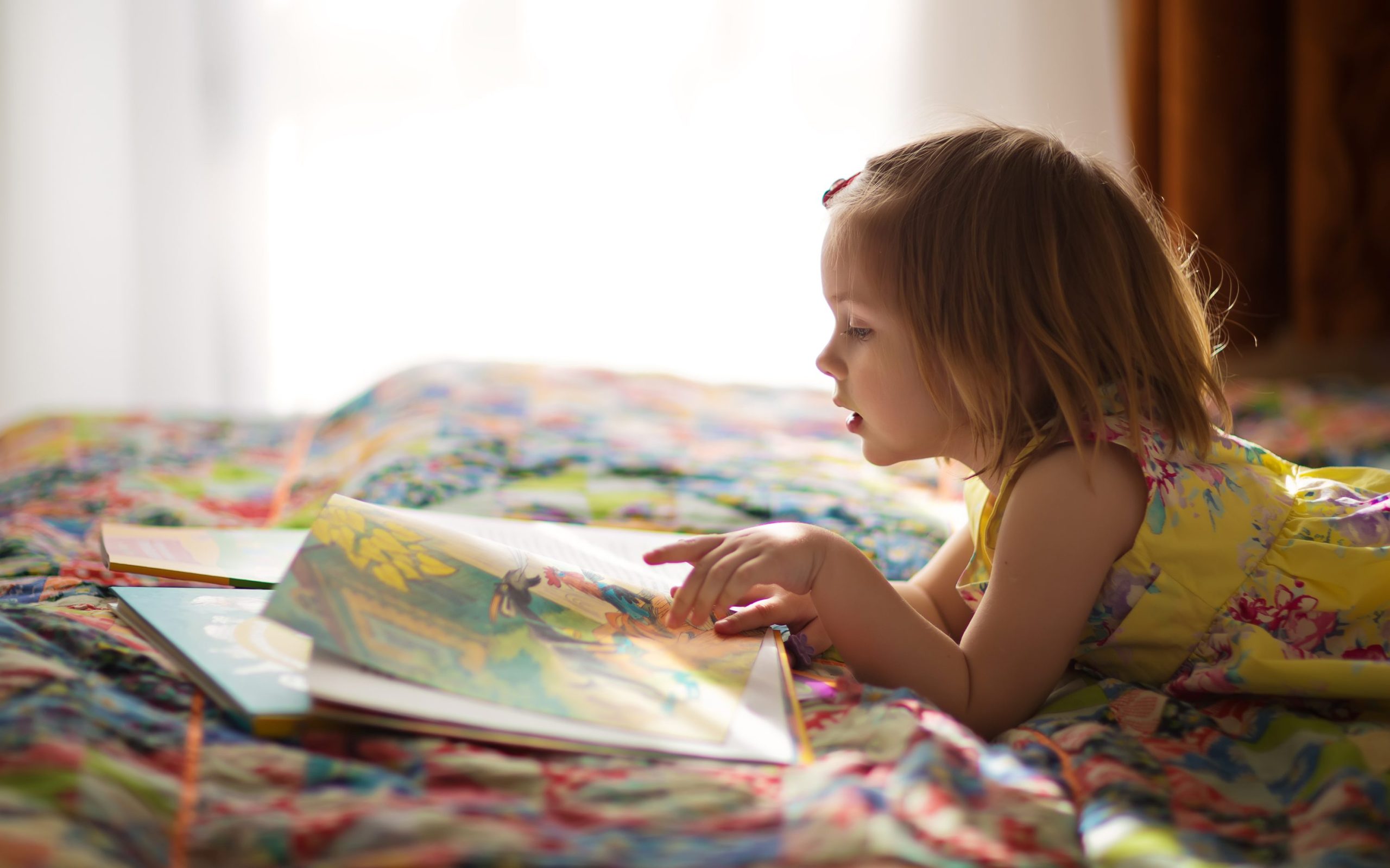 A photograph of a young girl lying on a bed reading a book.