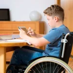 Photo of a boy in a wheelchair holding a tablet. He is sat at a table with books in front of him.