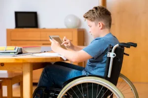 Photo of a boy in a wheelchair holding a tablet. He is sat at a table with books in front of him.