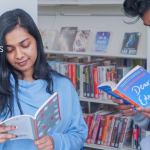 A woman in blue and a man in a grey hoodie stand reading in a library