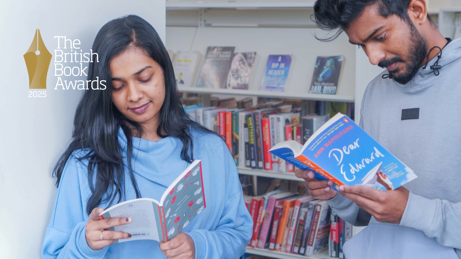 A woman in blue and a man in a grey hoodie stand reading in a library